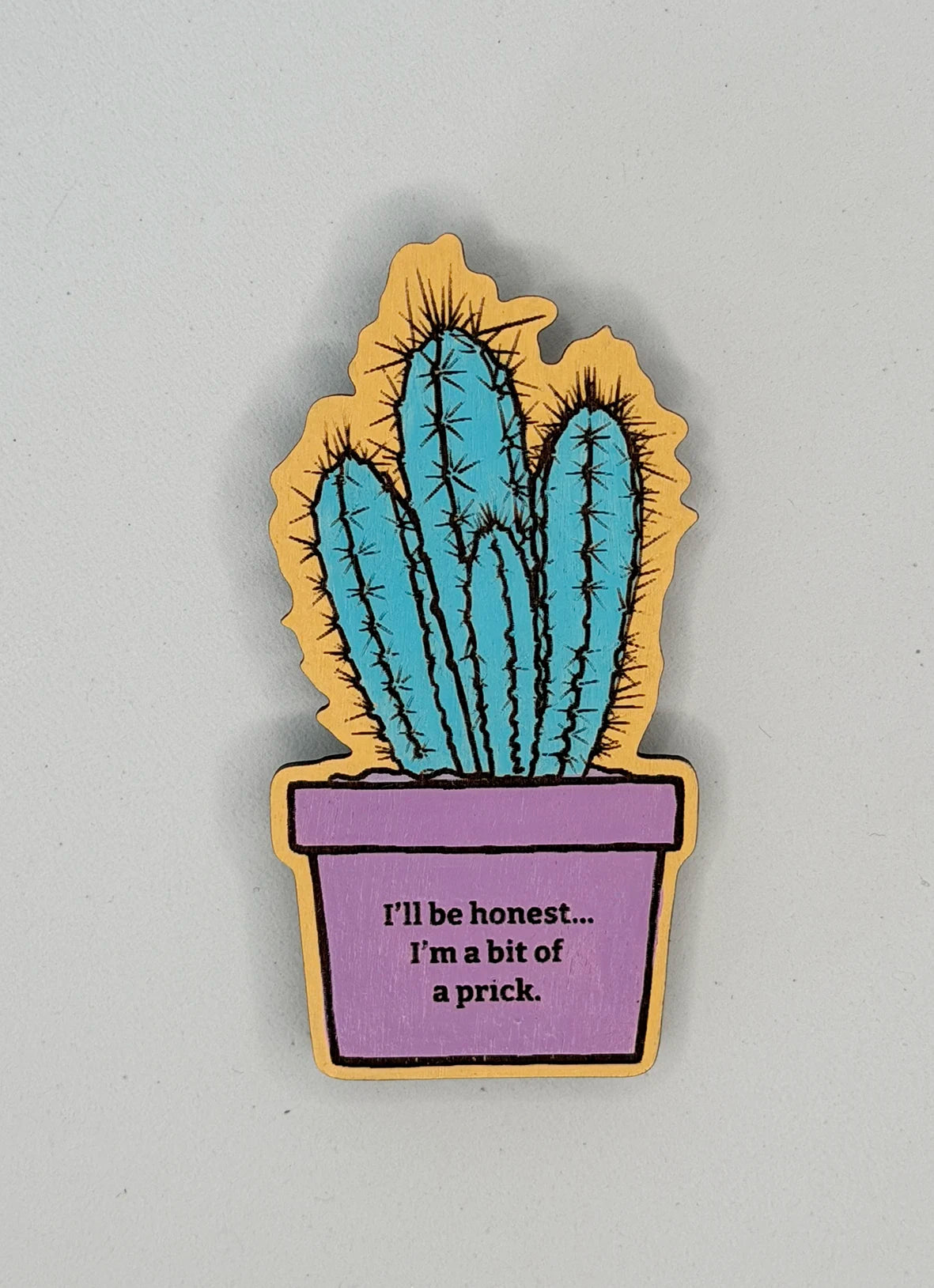 Original cactus illustration engraved onto wood and turned into magnet, The cactus has been hand-painted in turquoise and sits in a lilac plant pot. than plant sits on a light range background.