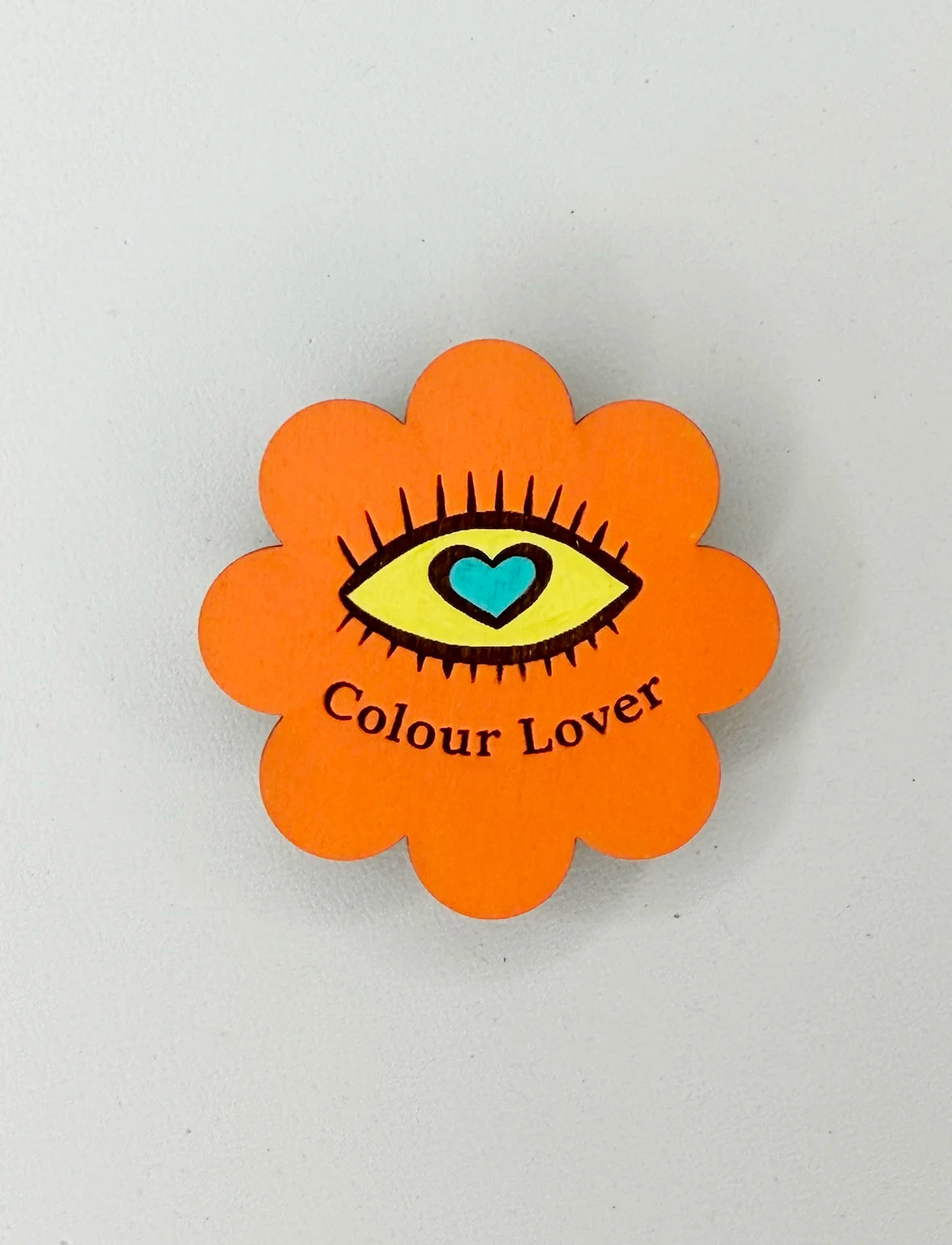 An orange flower shaped magnet with a yellow eye illustration with a turquoise love heart within. Text reads Colour Lover.