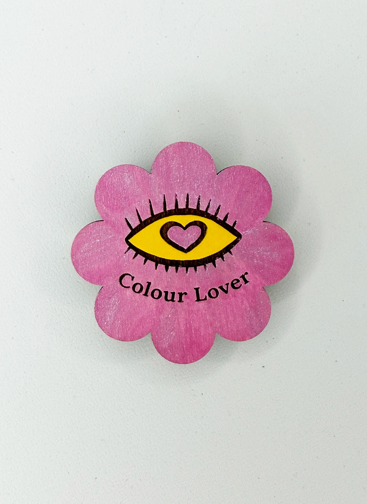 A pink flower-shaped wooden pin or badge with a yellow eye and heart design in the center, and the words 'Colour Lover' printed below, shown on a plain light background.