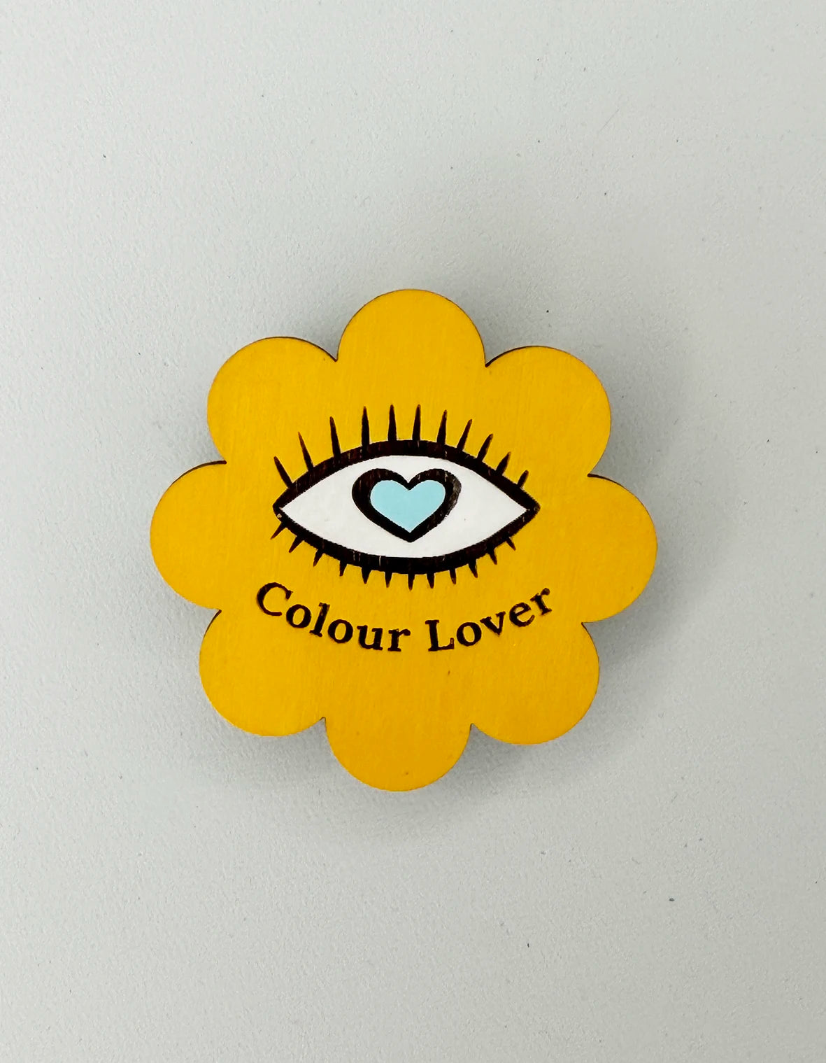 Flower shaped yellow magnet with an engraved bold eye illustration with a baby blue love heart iris within. Engraved text reads Colour Lover.